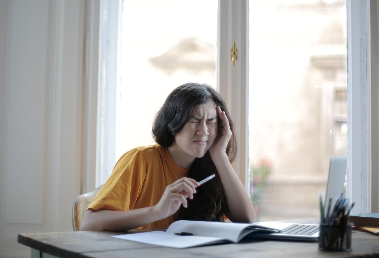 Estudiante sufriendo una migraña mientras intenta estudiar con su computadora portátil en casa.