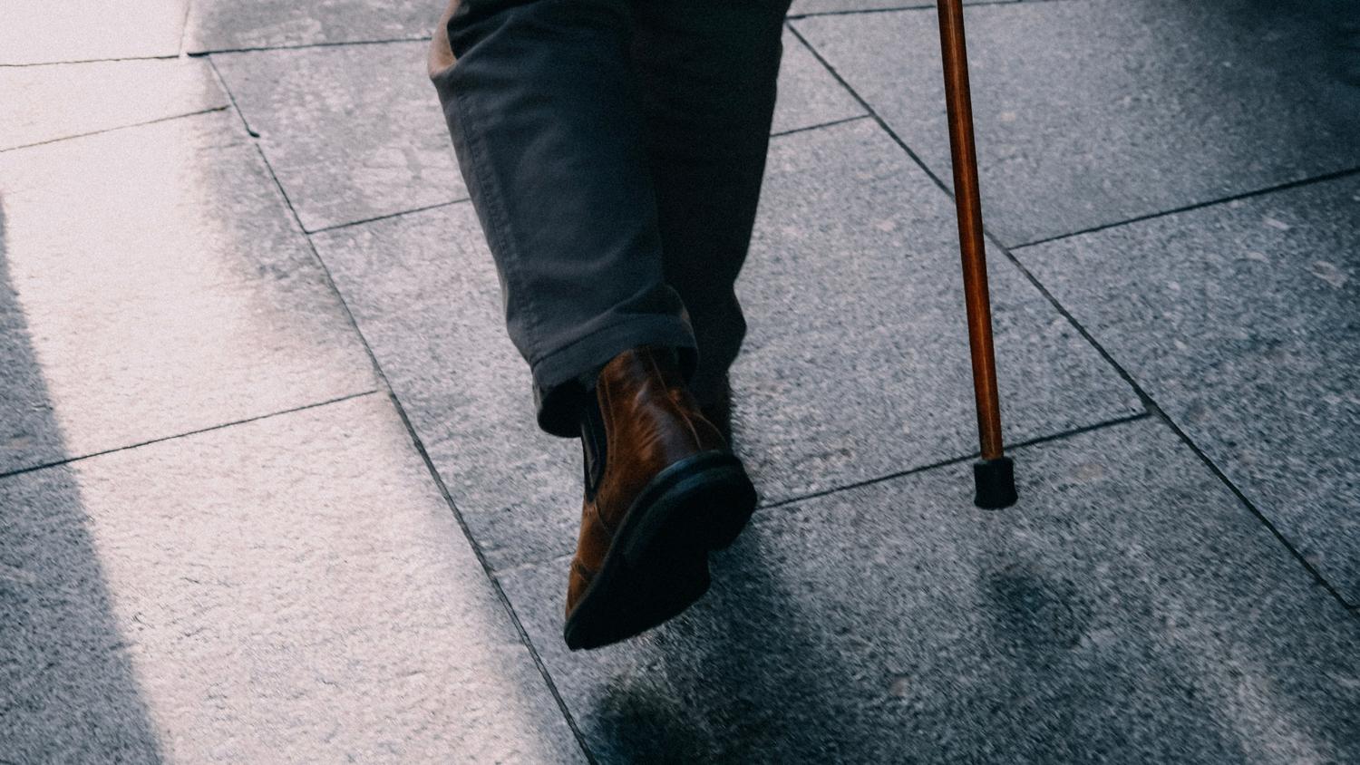 Detalhe dos sapatos e o bastão de uma pessoa idosa sobre o pavimento, focando nos elementos de mobilidade e estabilidade.