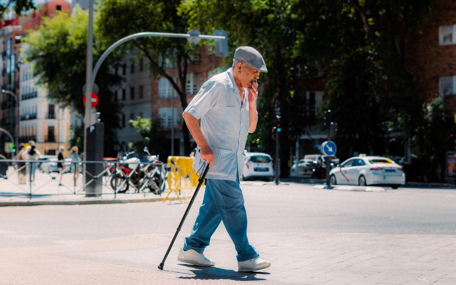 Um homem idoso usando um bastão para caminhar pela rua, representando o uso de dispositivos de apoio e o risco de quedas.