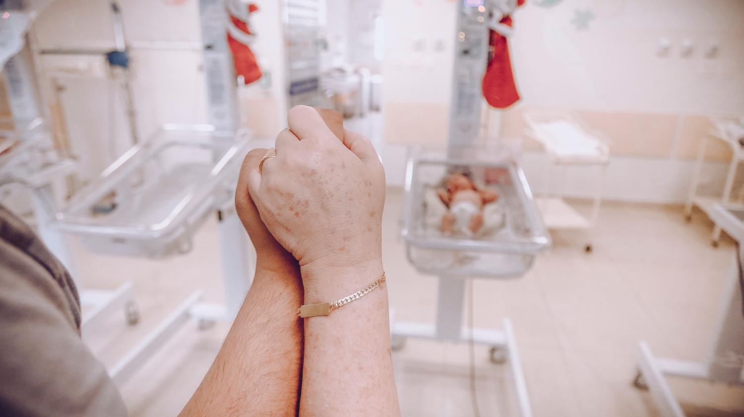 Manos de los padres entrelazadas en una guardería hospitalaria con recién nacidos al fondo, simbolizando el apoyo emocional.