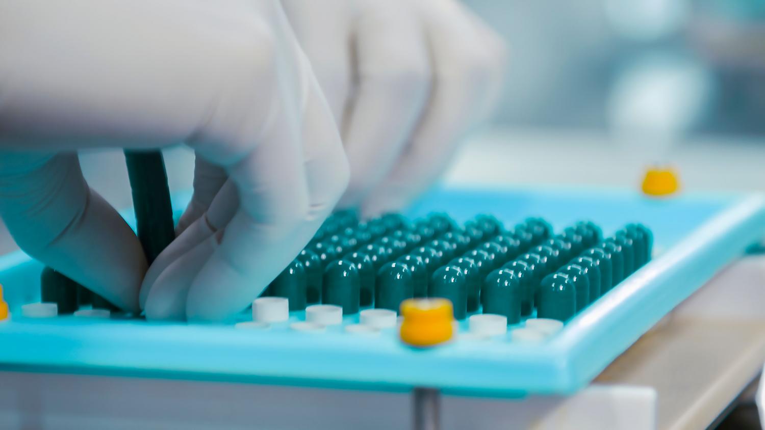 Manos con guantes manipulando cápsulas en un laboratorio farmacéutico, ilustrando la transición de fármaco a medicamento.