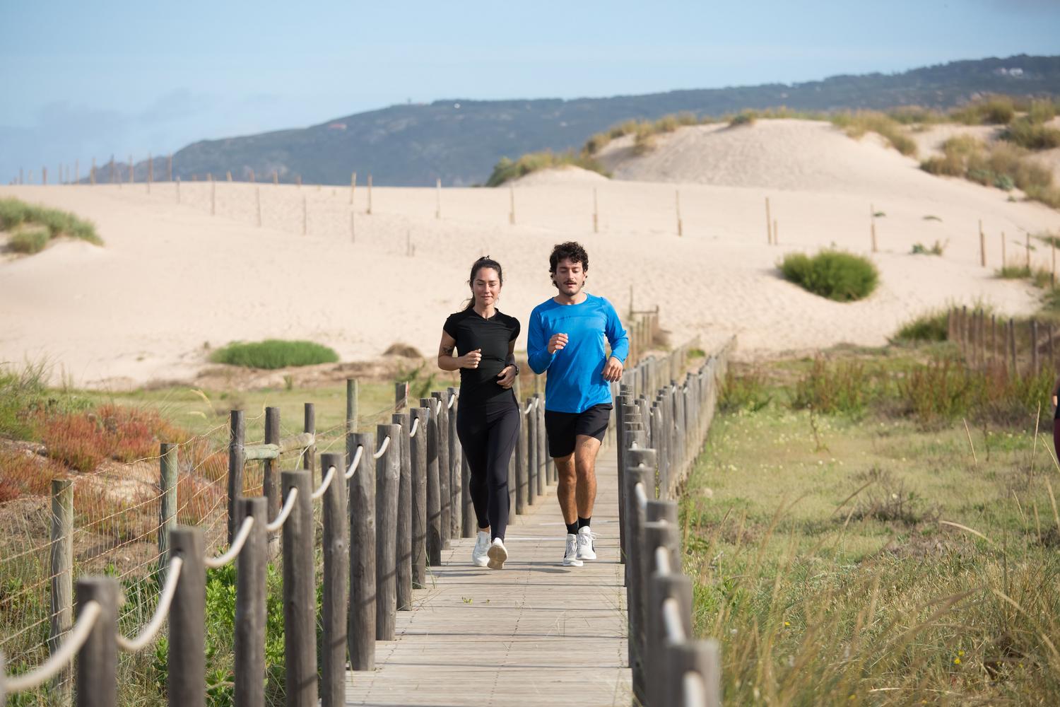 Hombre y mujer trotando por una pasarela de madera en un entorno de dunas costeras bajo el sol