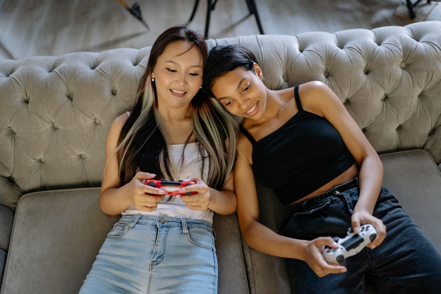 Duas mulheres jovens relaxadas jogando videogames juntas em um sofá, ilustrando o aspecto social e o combate à solidão.