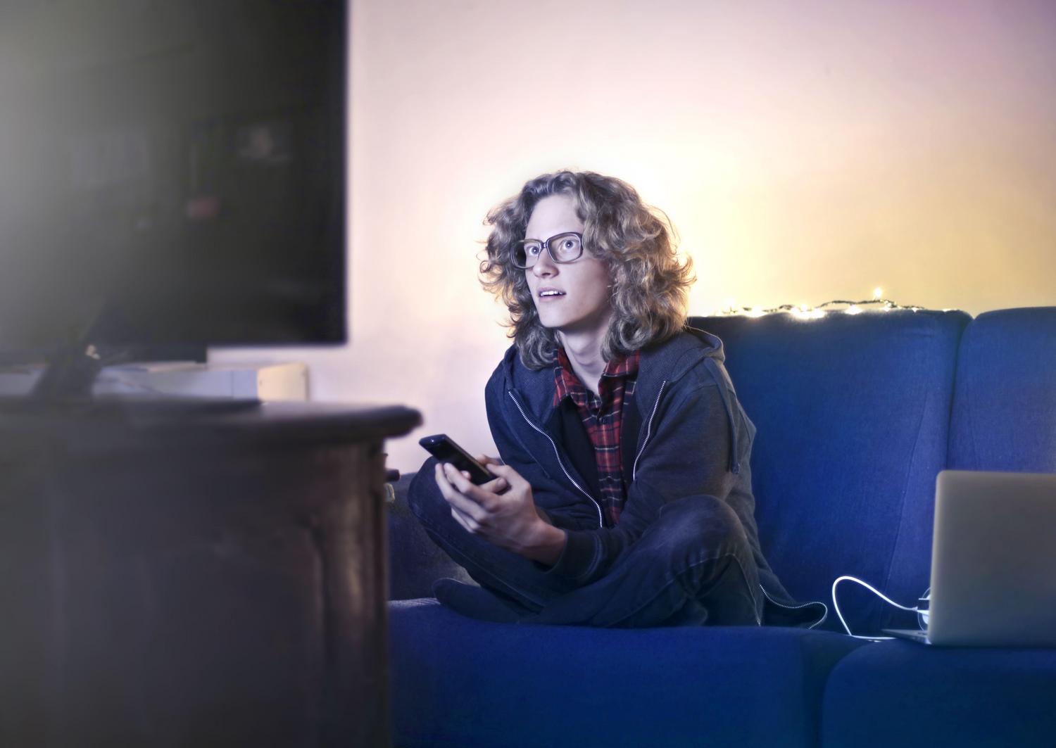 Adolescente utilizando su teléfono móvil mientras ve la televisión, ilustrando la sobreexposición a múltiples pantallas y el consumo simultáneo de redes sociales.