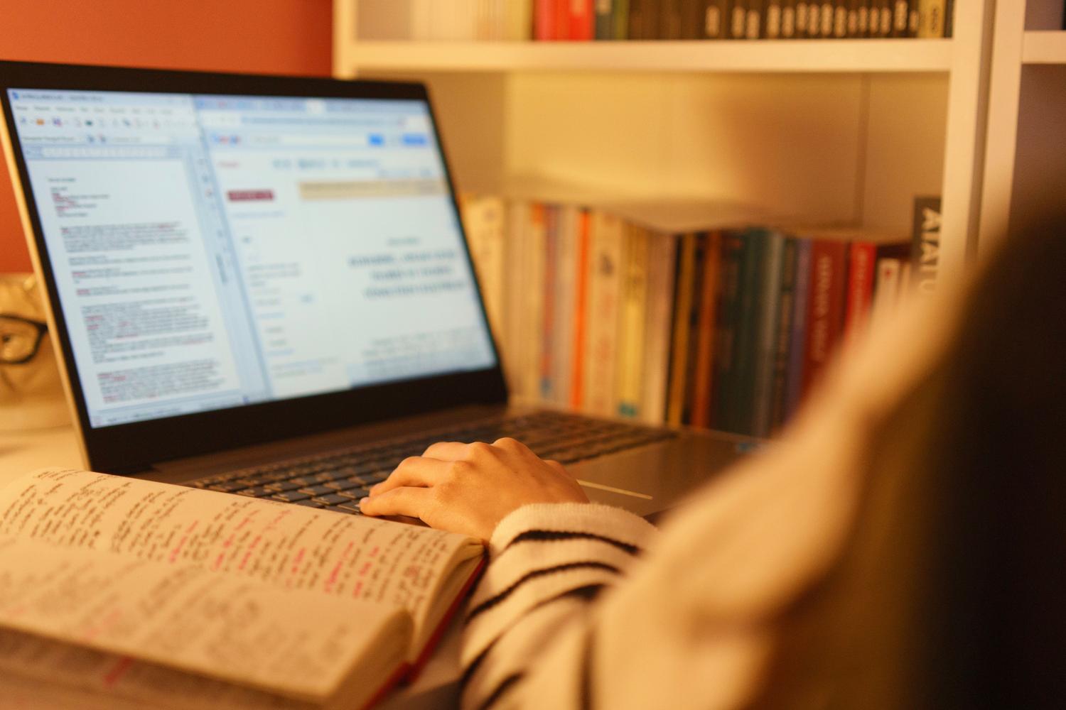 Estudiante redactando un trabajo académico en casa utilizando una laptop y libros de referencia.