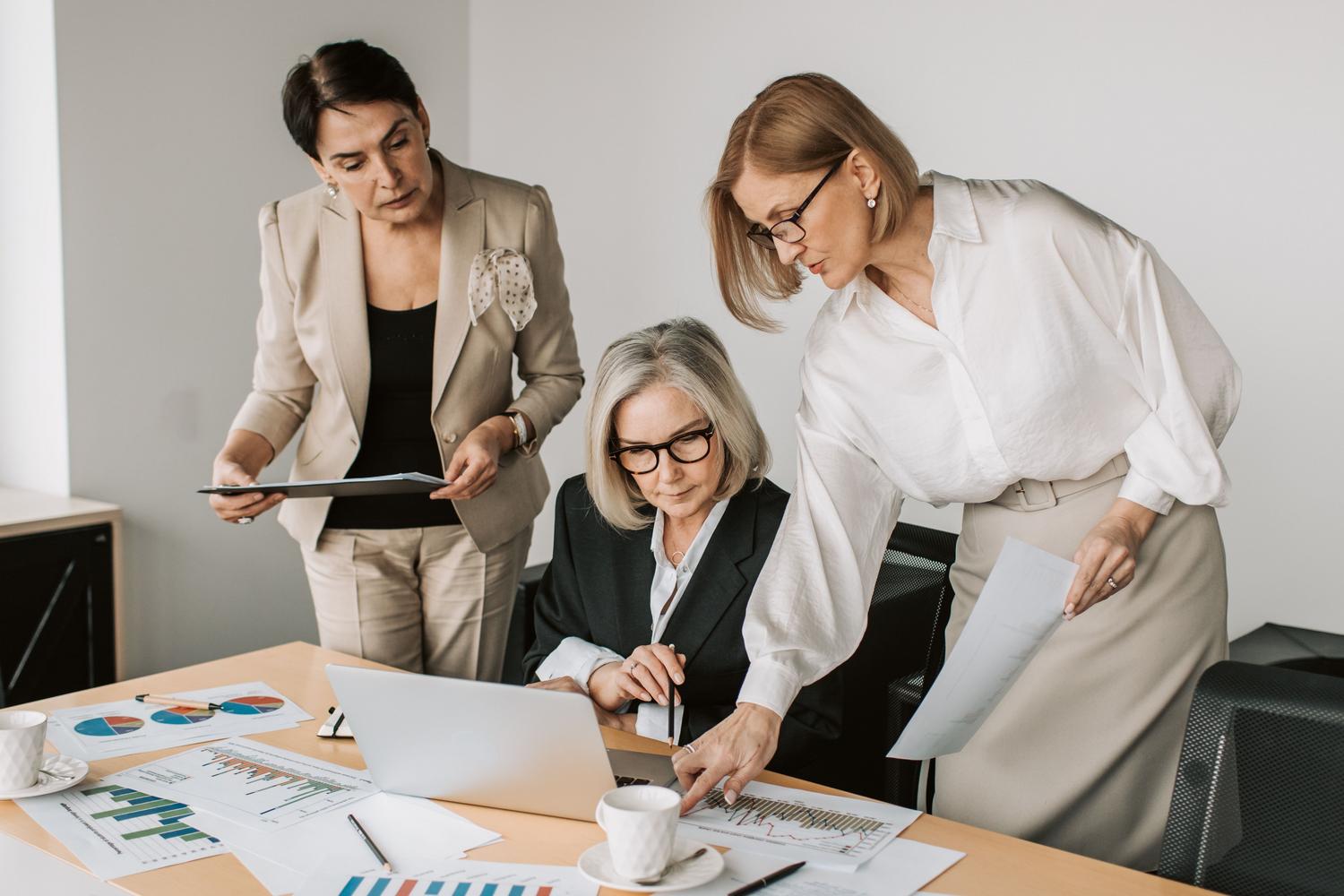 Tres mujeres de alta dirección colaborando con un ordenador portátil y reportes financieros en una oficina luminosa, mostrando la parte operativa y la integración de procesos de la gestión senior.