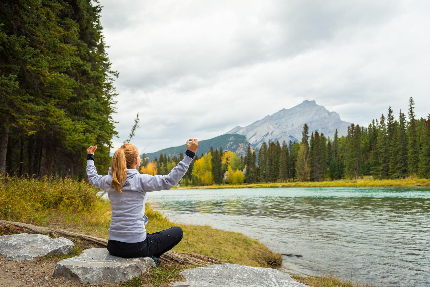 Mujer meditando en calma junto a un lago rodeado de montañas, representando la recuperación de la salud mental.