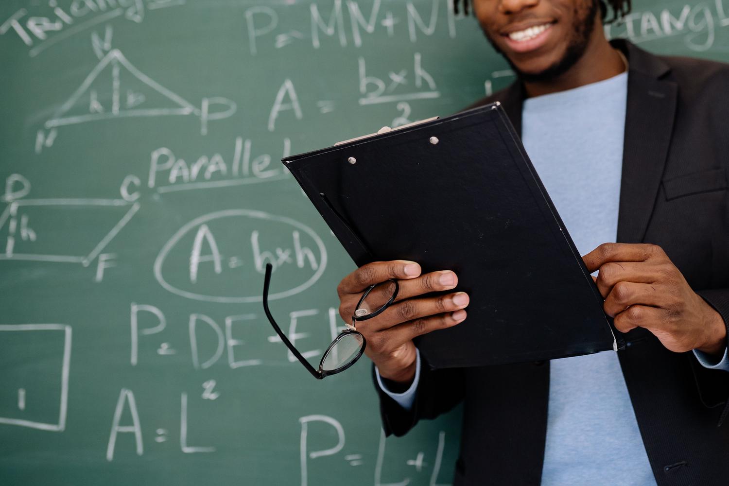 Profesor de matemáticas sonriente y accesible frente a una pizarra con fórmulas geométricas.