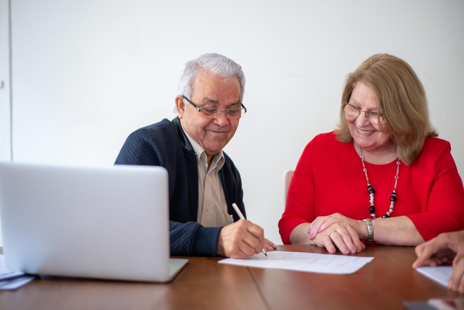Pareja senior firmando documentos oficiales y utilizando un ordenador portátil para gestionar su pensión