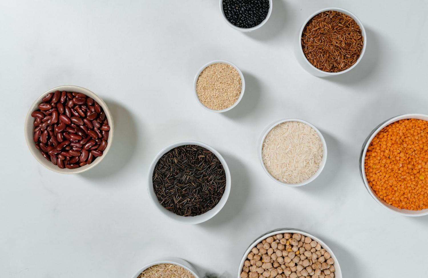 Variedad de legumbres y granos integrales en cuencos sobre una superficie de mármol blanco, representando la diversidad de proteínas y fibras vegetales.