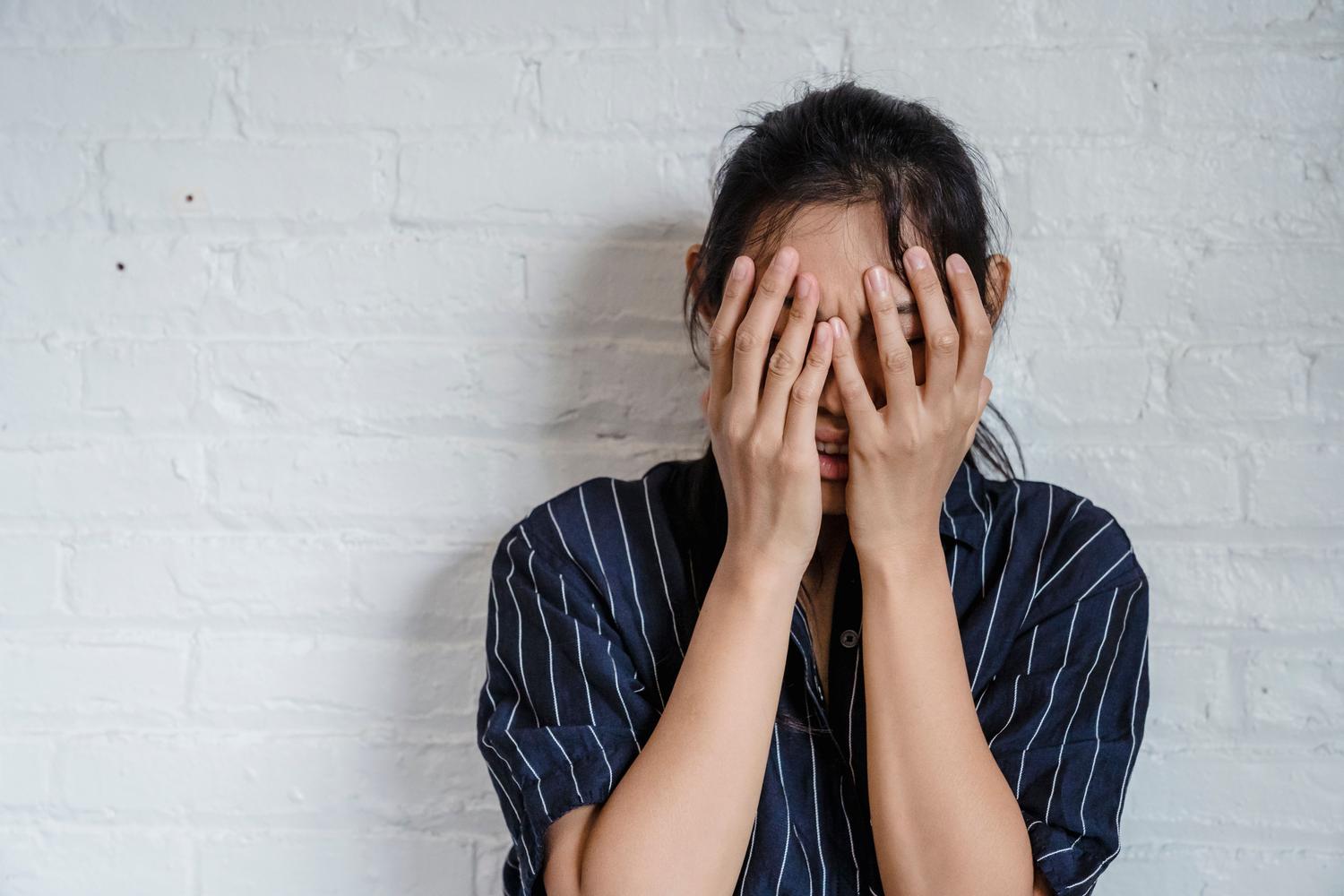 Mujer joven expresando estrés y ansiedad cubriéndose el rostro con las manos contra una pared blanca