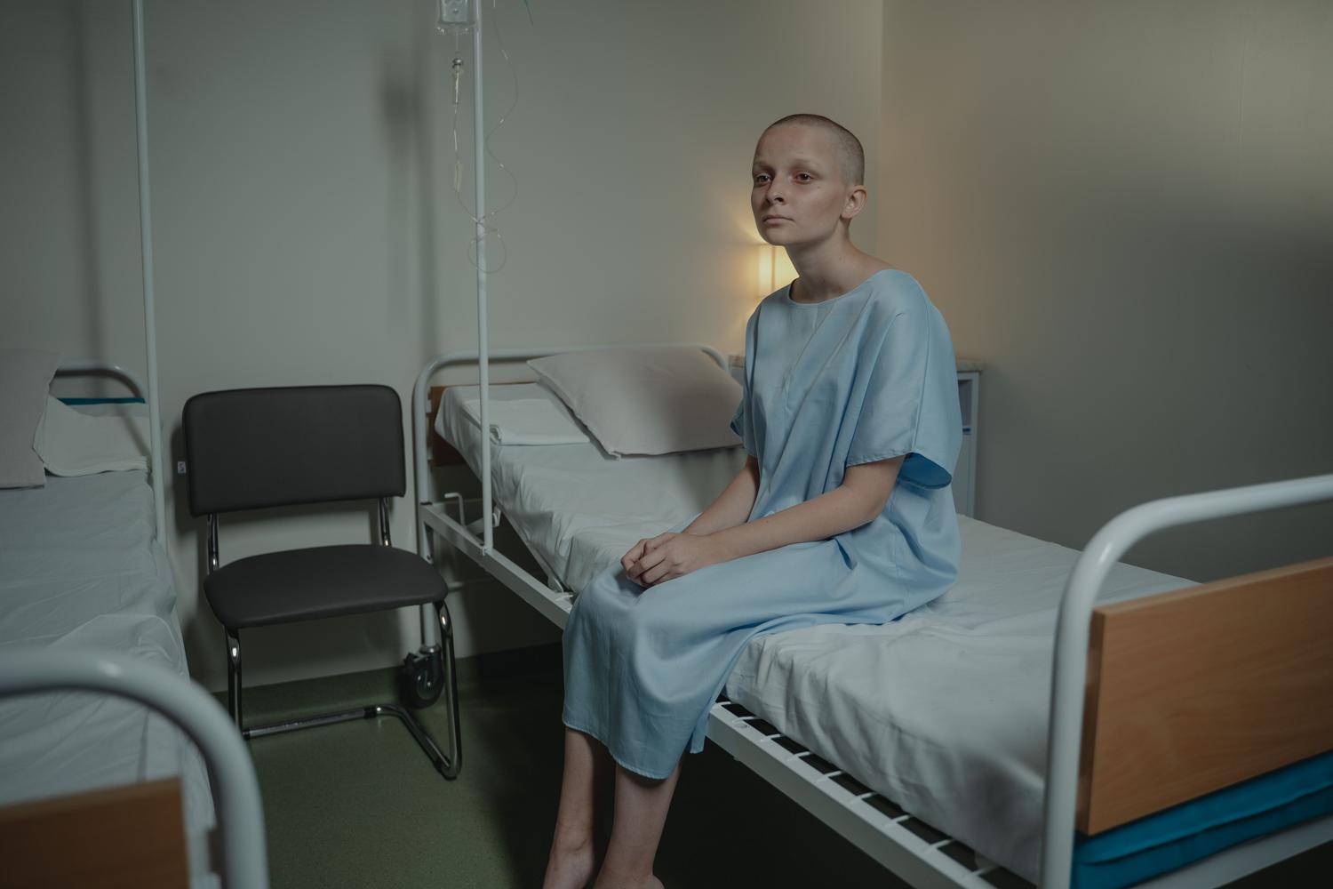 Paciente joven calva sentada en una cama de hospital con expresión reflexiva y sombría, representando el impacto emocional del diagnóstico.