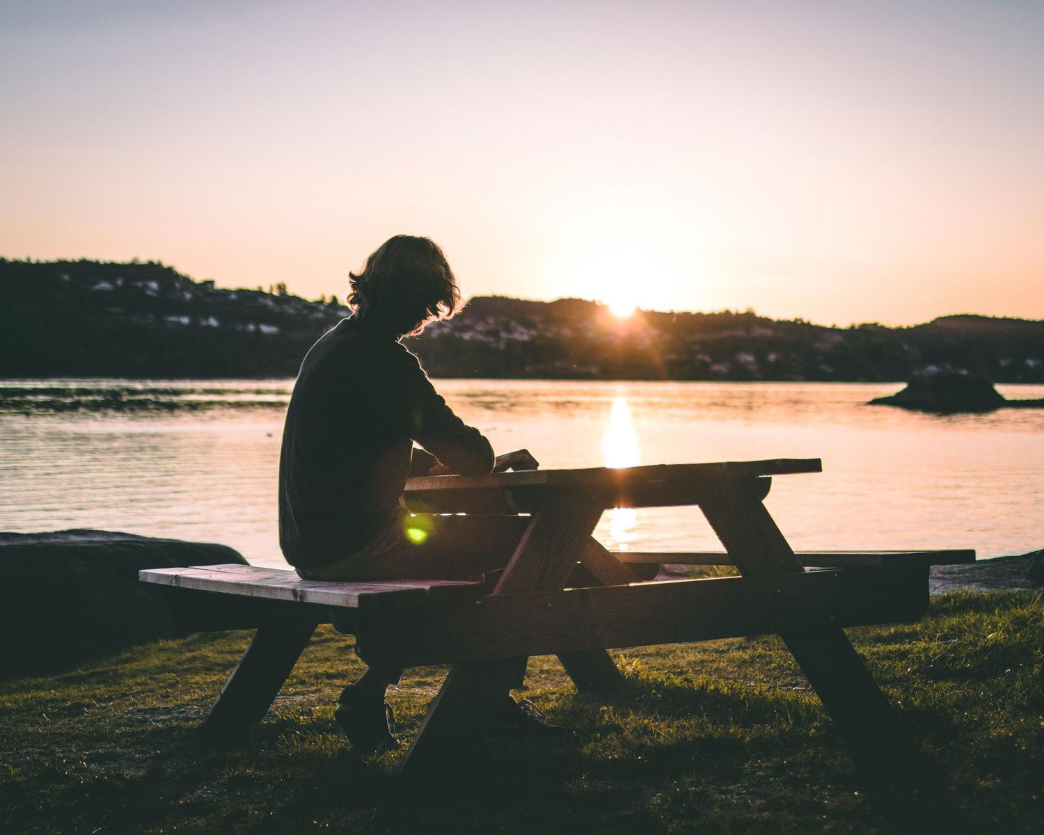 Hombre contemplativo sentado en un banco frente a un lago tranquilo al atardecer, ilustrando la reflexión interna.