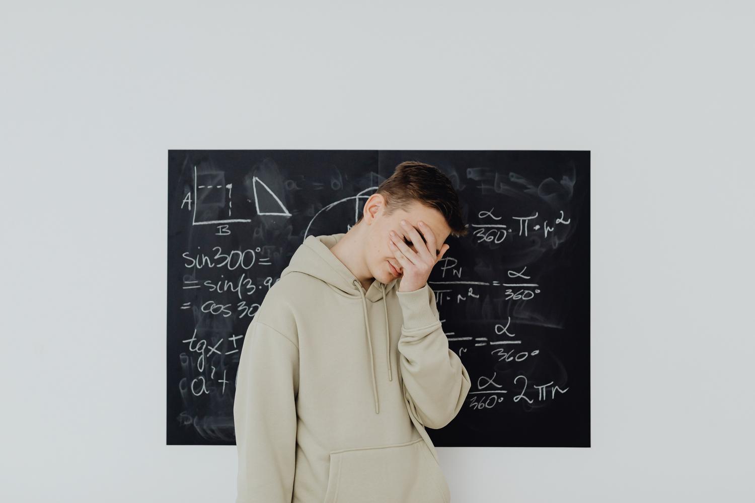 Estudiante adolescente con sudadera cubriéndose el rostro en señal de frustración frente a una pizarra de matemáticas.